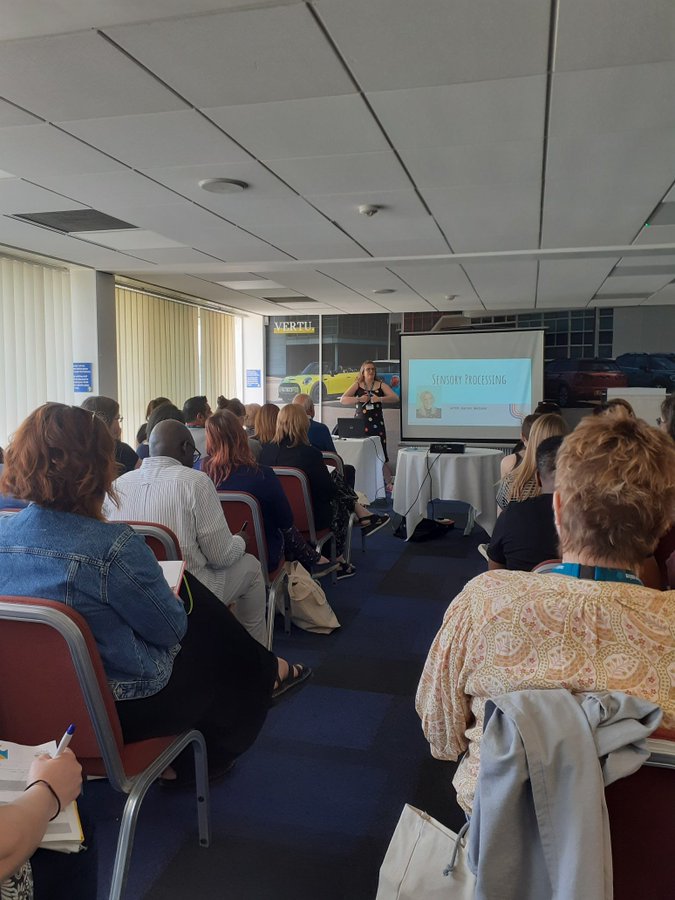 Karen is presenting a workshop to a room of seated participants. The screen behind her says Sensory Processing.