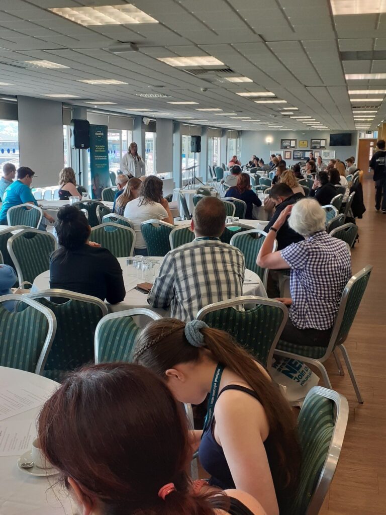 Circular tables surrounded by seated people, listening to the keynote speaker at the Send Help conference.